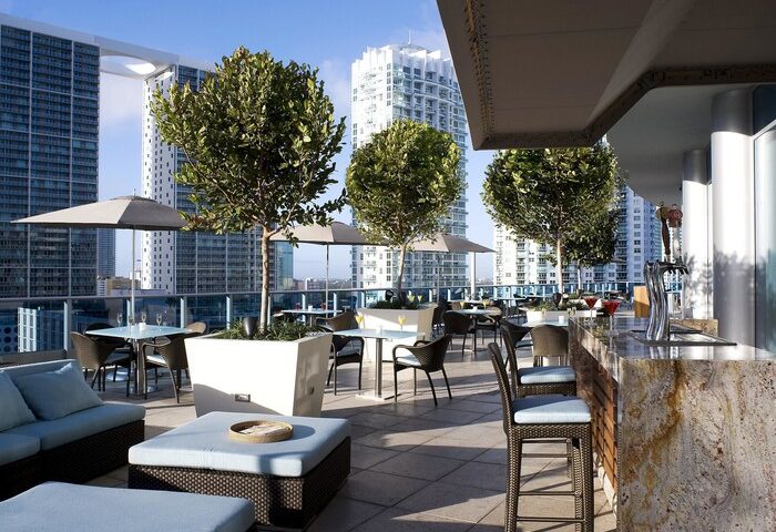 Kimpton EPIC Hotel's rooftop bar. A gay-friendly hotel with city views, tables, chairs, umbrellas, trees, and a bar area. Miami travel.