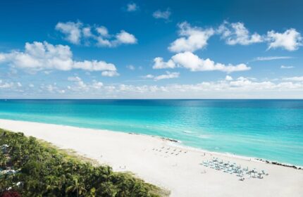 Miami Beach EDITION gay-friendly hotel view. Turquoise ocean, white sand beach, blue sky with clouds. Palm trees line the coast.