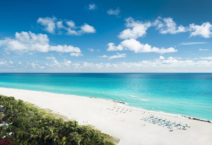 Miami Beach EDITION gay-friendly hotel view. Turquoise ocean, white sand beach, blue sky with clouds. Palm trees line the coast.