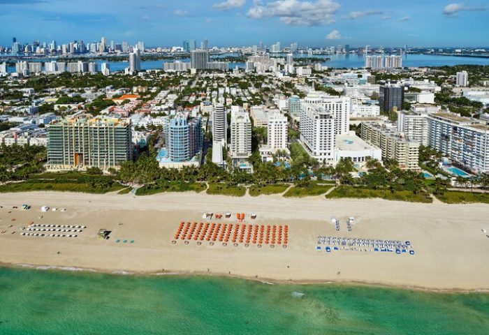 Aerial view of Royal Palm South Beach Miami, a gay-friendly hotel. Beach umbrellas line the shore. Skyline in background.