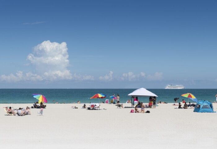 Beach scene near President Hotel, a gay-friendly hotel. People relax under colorful umbrellas on the white sand. Cruise ship in the distance.