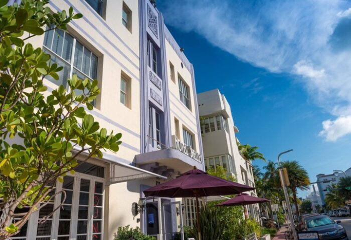 Hotel Shelley, a gay-friendly hotel. Art Deco architecture with purple accents. Sunny day, palm trees, and outdoor seating. Great for gay travel.