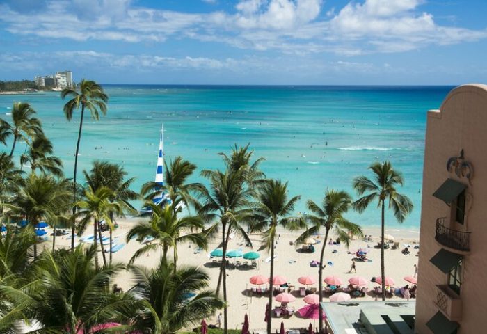Waikiki beach view from The Royal Hawaiian, a Luxury Collection Resort. Gay-friendly hotel with palm trees, pink umbrellas, and turquoise ocean.