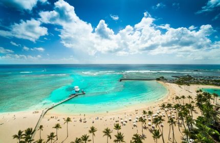 Aerial view of Waikiki Beach at Hilton Hawaiian Village, a gay-friendly hotel. Turquoise water, palm trees, and a pier with a boat.