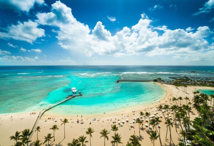 Aerial view of Waikiki Beach at Hilton Hawaiian Village, a gay-friendly hotel. Turquoise water, palm trees, and a pier with a boat.