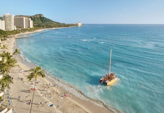 Outrigger Waikiki Beach Resort view. Gay-friendly hotel on a sandy beach with palm trees, Diamond Head, and a catamaran. Waikiki vacation.