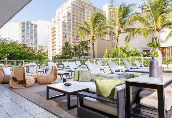 Hilton Garden Inn Waikiki Beach gay-friendly hotel. Outdoor lounge area with chairs, tables, and lounge chairs. Palm trees and buildings in background.