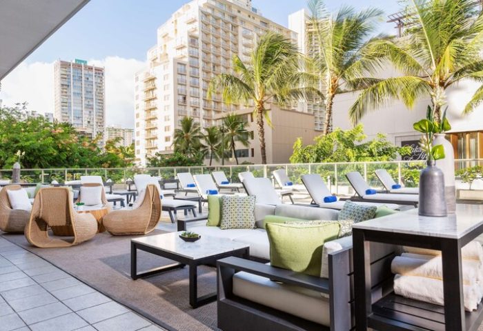 Hilton Garden Inn Waikiki Beach gay-friendly hotel. Outdoor lounge area with chairs, tables, and lounge chairs. Palm trees and buildings in background.