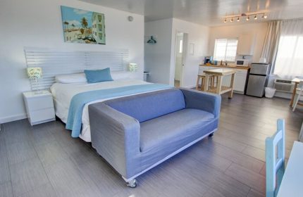 Gay-friendly hotel room with a bed, couch, and kitchenette. A painting of a beach scene hangs above the bed. Modern, clean design. Snooze.