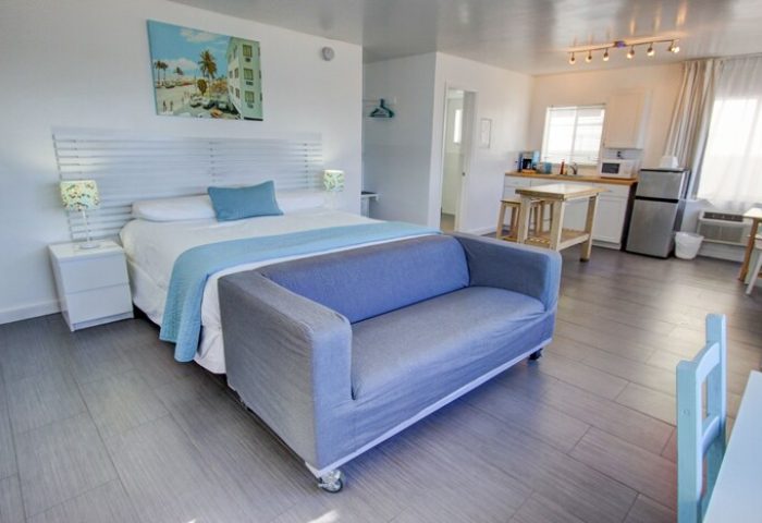 Gay-friendly hotel room with a bed, couch, and kitchenette. A painting of a beach scene hangs above the bed. Modern, clean design. Snooze.