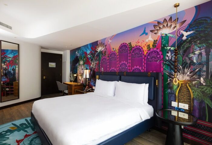 Hotel Indigo Dubai Downtown gay-friendly room. King bed with white linens, colorful mural, desk, mirror, and side table. Modern decor.