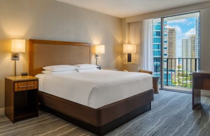 Hyatt Regency Miami hotel room. Gay-friendly hotel with balcony view of Miami skyline. Bedroom with king bed, nightstand, and modern decor.