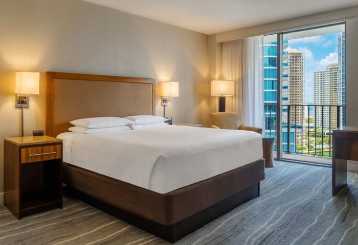 Hyatt Regency Miami hotel room. Gay-friendly hotel with balcony view of Miami skyline. Bedroom with king bed, nightstand, and modern decor.