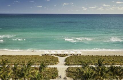 Four Seasons Hotel at The Surf Club beach view. Gay-friendly hotel with white umbrellas, turquoise ocean, palm trees, and cyclists. Ideal gay travel spot.