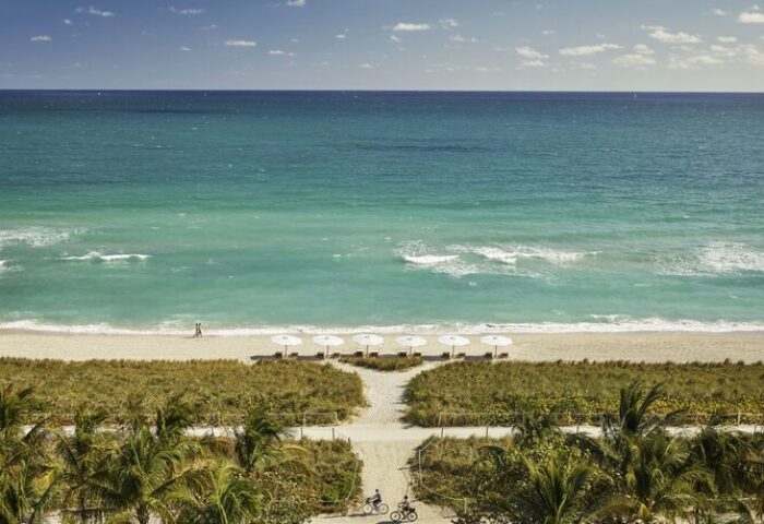 Four Seasons Hotel at The Surf Club beach view. Gay-friendly hotel with white umbrellas, turquoise ocean, palm trees, and cyclists. Ideal gay travel spot.