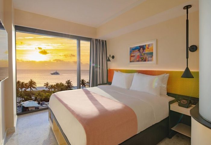 Moxy Miami South Beach gay-friendly hotel room with ocean view. Sunset over the sea, palm trees, and a yacht. Modern, stylish decor.