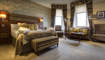 Crieff Hydro gay-friendly hotel room. Bedroom with a king-size bed, striped bench, patterned wallpaper, curtains, sofa, and chairs.