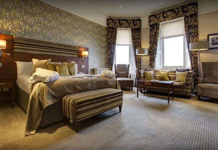 Crieff Hydro gay-friendly hotel room. Bedroom with a king-size bed, striped bench, patterned wallpaper, curtains, sofa, and chairs.