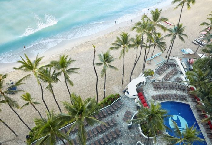 Aerial view of Outrigger Waikiki Beach Resort, a gay-friendly hotel. Palm trees, pool, lounge chairs, & beach create a tropical paradise. Perfect for gay travel.