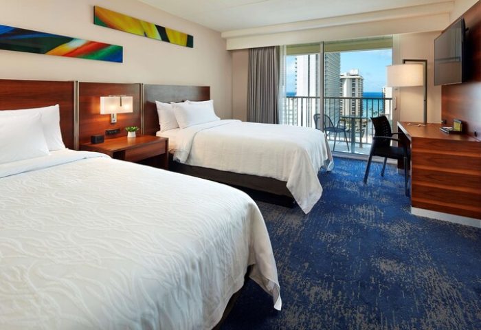 Hilton Garden Inn Waikiki Beach gay-friendly hotel room. Two beds, balcony with ocean view. Modern decor, desk, and blue carpet.