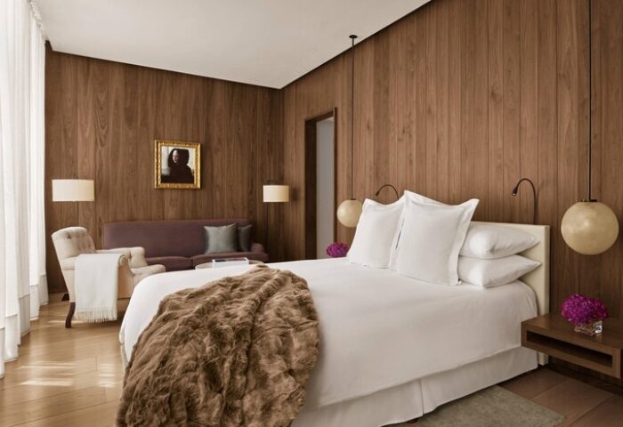 The London EDITION hotel room. Gay-friendly hotel with wood paneling, white linens, and a brown faux fur throw. Sofa and lamps visible.