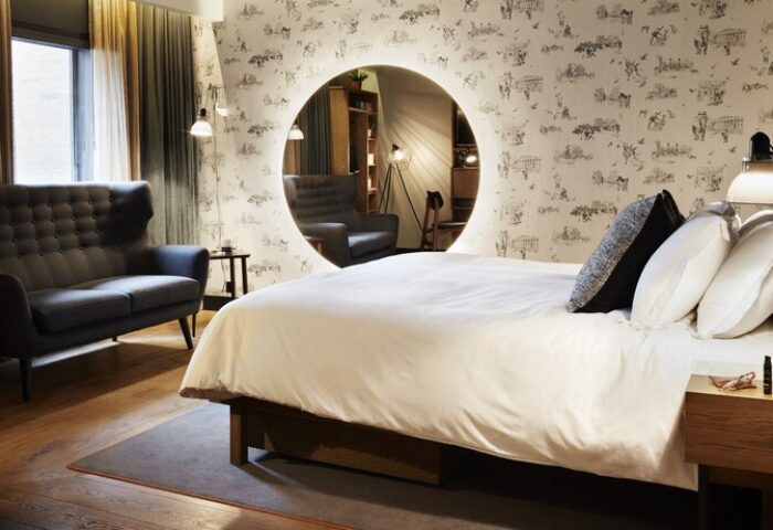 The Hoxton, Holborn gay-friendly hotel room. Double bed with white linen, a sofa, and a round mirror on a patterned wall. Modern design.