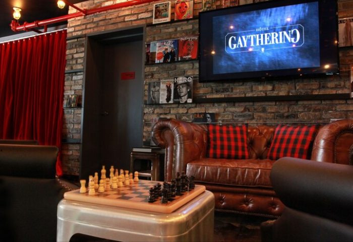 Interior of the Gaythering, a gay-friendly hotel. Chessboard, leather sofa, and TV with the hotel's logo. Brick wall and red accents.