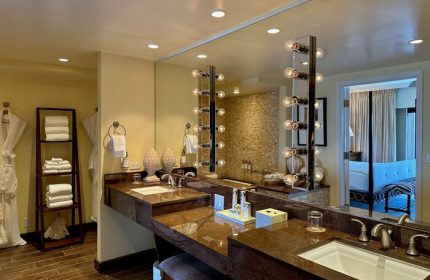 Kahala Hotel and Resort gay-friendly bathroom with double vanity, lighted mirror, towels, robes, and a glimpse into the bedroom. Luxury hotel.