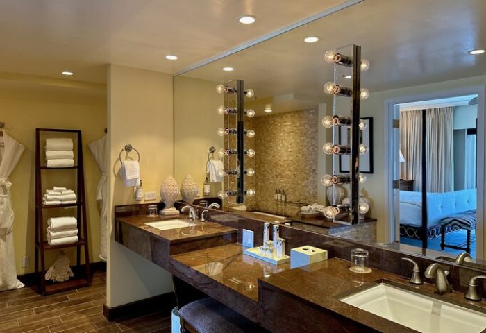 Kahala Hotel and Resort gay-friendly bathroom with double vanity, lighted mirror, towels, robes, and a glimpse into the bedroom. Luxury hotel.
