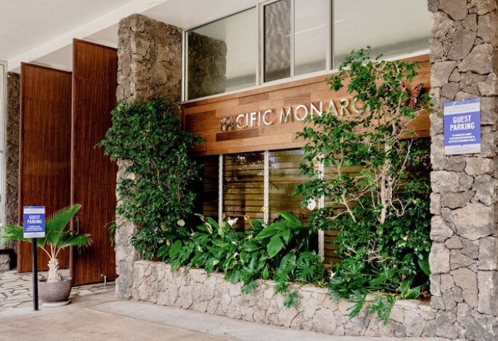 Pacific Monarch Hotel entrance. Gay-friendly hotel with stone facade, wood accents, and lush greenery. Guest parking sign visible.