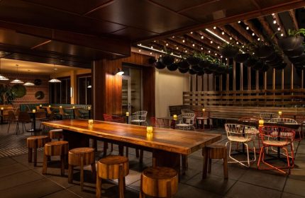 The Laylow, Autograph Collection gay-friendly hotel outdoor dining. Long wooden table with stools, string lights, and hanging plants create a warm ambiance.