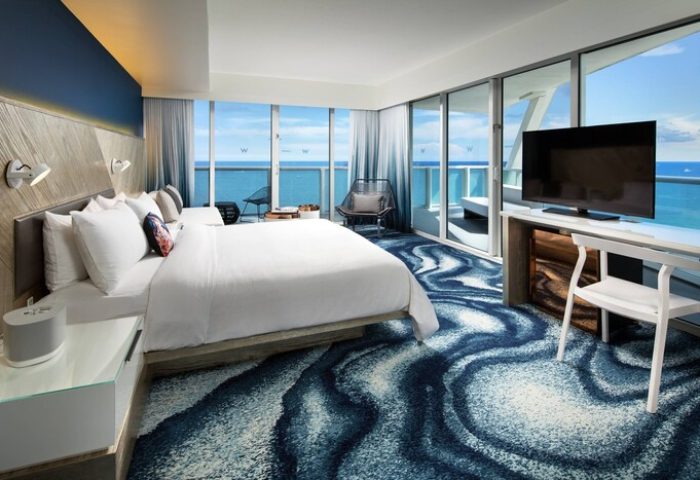 W Fort Lauderdale gay-friendly hotel room with ocean view. Modern design, king bed, balcony, and blue wave-patterned carpet. Luxury travel.