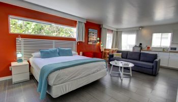 Gay-friendly hotel room with bed, seating area, and kitchenette. Modern design, orange accent wall. Ideal for gay men's travel. Snooze in style.