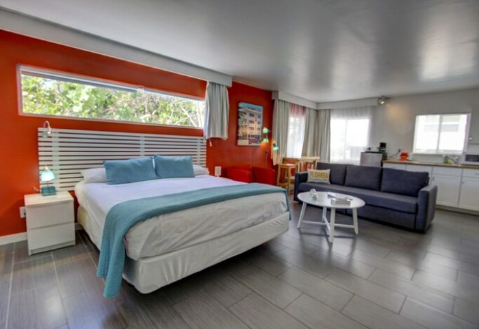 Gay-friendly hotel room with bed, seating area, and kitchenette. Modern design, orange accent wall. Ideal for gay men's travel. Snooze in style.