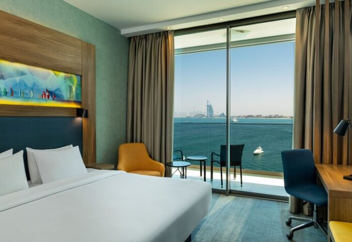 Aloft Palm Jumeirah gay-friendly hotel room. Balcony view of Dubai skyline and ocean. Modern design, bed, desk, chair. Travel destination.
