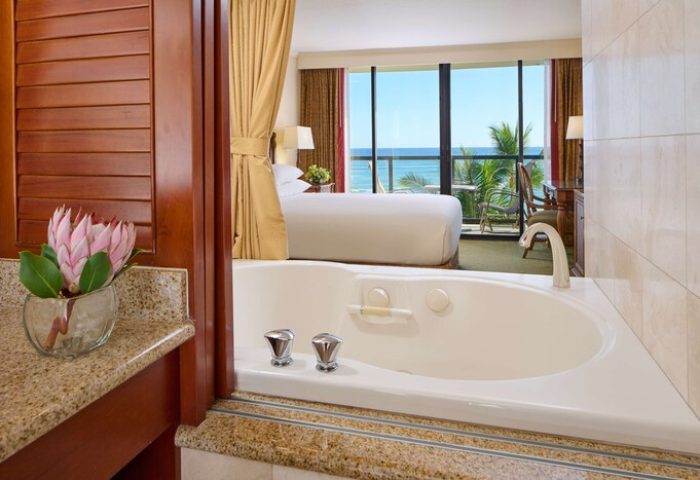 Outrigger Waikiki Beach Resort gay-friendly hotel room view. Bathtub in foreground, bedroom with ocean view, balcony, and palm trees in the background.