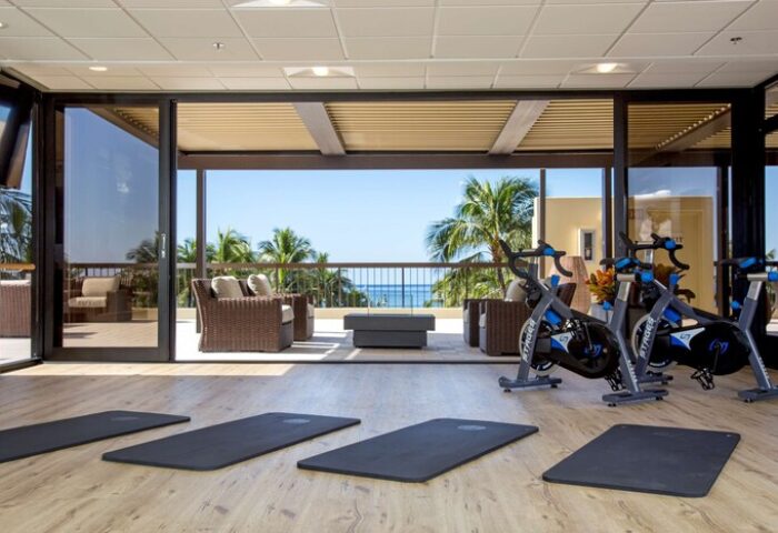 Aston Waikiki Beach Tower gym with ocean view. Gay-friendly hotel fitness center with exercise bikes and yoga mats. Palm trees visible.