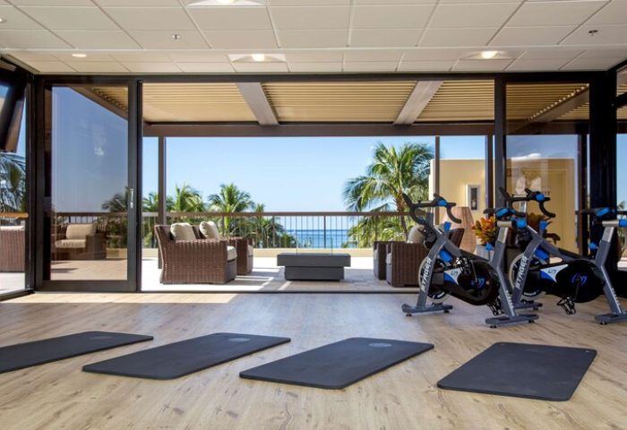 Aston Waikiki Beach Tower gym with ocean view. Gay-friendly hotel fitness center with exercise bikes and yoga mats. Palm trees visible.