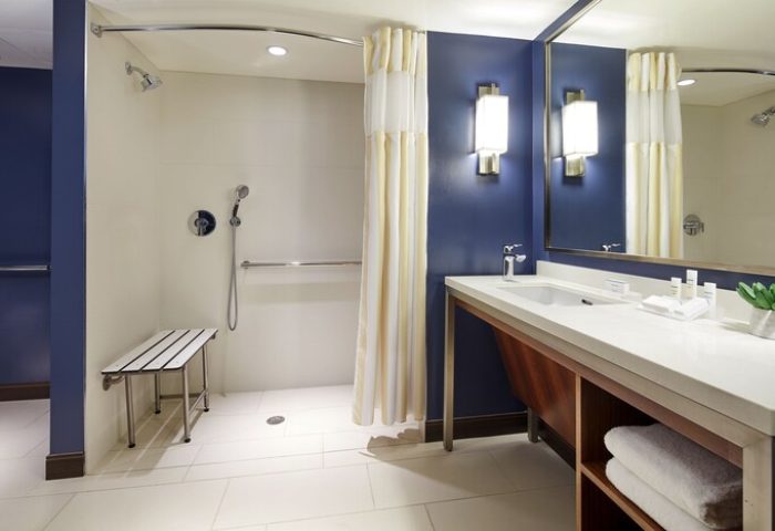 Hilton Garden Inn Waikiki Beach gay-friendly hotel bathroom. Accessible shower with bench, grab bar, and handheld shower head. Vanity with sink.