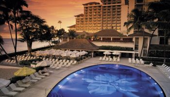 Halekulani Hotel pool at sunset. Gay-friendly hotel with ocean view, lounge chairs, and palm trees. Luxury travel for gay men in Honolulu.