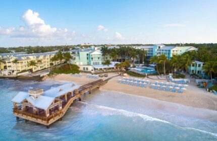 The Reach Key West, Curio Collection by Hilton, a gay-friendly hotel. Aerial view of beach, pier, and hotel buildings. Palm trees and blue skies.
