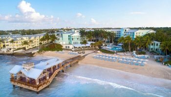 The Reach Key West, Curio Collection by Hilton, a gay-friendly hotel. Aerial view of beach, pier, and hotel buildings. Palm trees and blue skies.