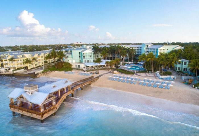 The Reach Key West, Curio Collection by Hilton, a gay-friendly hotel. Aerial view of beach, pier, and hotel buildings. Palm trees and blue skies.