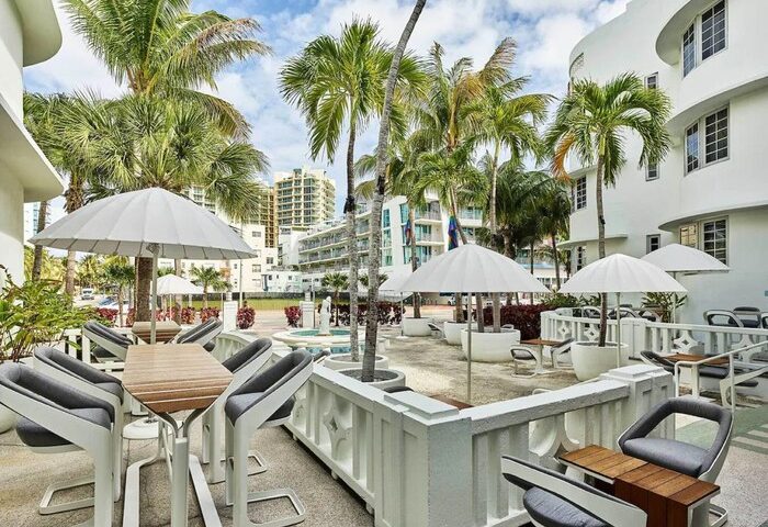 Axel Beach Miami gay-friendly hotel outdoor seating area. Tables with umbrellas, chairs, palm trees, and white building. Gay travel destination.