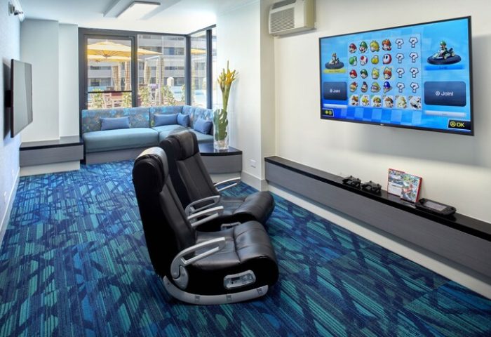 Holiday Inn Express Waikiki gay-friendly hotel game room. Two black gaming chairs face a TV with Mario Kart. Blue patterned carpet. Sofa seating.