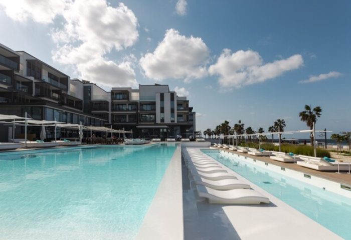 Nikki Beach Resort & Spa Dubai pool view. Gay-friendly hotel with modern architecture, sun loungers, palm trees, and blue sky. Ideal for gay travel.