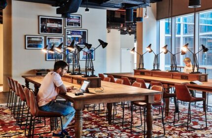 25hours Hotel One Central co-working space. Gay-friendly hotel with long tables, chairs, and lamps. Men work on laptops in a stylish setting.