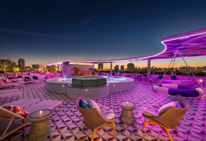Rooftop pool and lounge at Moxy Miami South Beach, a gay-friendly hotel. Purple lighting, city skyline, and stylish decor create a vibrant atmosphere.