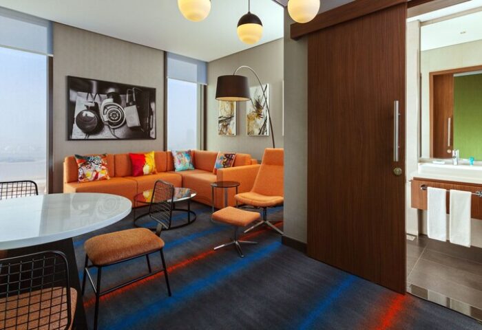 Aloft Me'aisam, Dubai: Gay-friendly hotel room with orange couch, chairs, and modern decor. Bathroom visible. A comfortable space for gay travelers.
