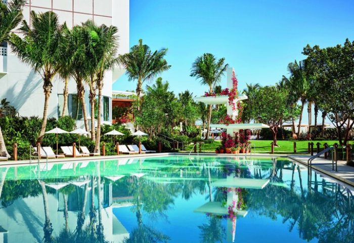 Miami Beach EDITION's pool. Gay-friendly hotel with palm trees, lounge chairs, and a serene pool reflecting the blue sky. A tropical paradise.